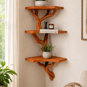 May include: A three-tier corner wall shelf featuring a brown wood-toned finish with supports sculpted to resemble tree branches. The shelves hold a small potted plant, a black cat figurine, a candle, and two books titled Kinfolk and Live Beautifully.