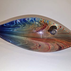 May include: A glass fish sculpture featuring a smooth, rounded body with a swirling pattern of deep blue, amber, and brown tones. The piece has a pointed head, a small circular eye detail, and a flared tail, showcasing a polished finish.