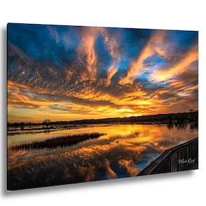 May include: A landscape photograph of a sunset over a wetland area with vibrant orange and blue clouds reflecting in the water. A dark wooden railing is visible in the bottom right corner, and the signature Mark Reid is printed in the corner.