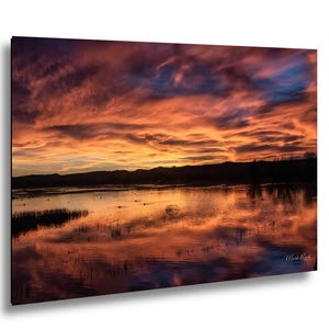 May include: A rectangular wall art print featuring a vibrant sunset over a calm body of water. The sky is filled with dramatic orange, red, and purple clouds reflecting on the water surface. Dark silhouettes of distant hills and reeds are visible.