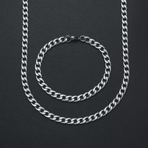 Może przedstawiać: Srebrzysty naszyjnik typu pancerka oraz pasująca okrągła bransoletka typu pancerka. Oba elementy posiadają zazębiające się, płaskie, owalne ogniwa z polerowanym metalicznym wykończeniem, tworząc jednolity i spójny wzór ogniw w całym zestawie.