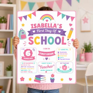 以下が含まれることがあります： 虹、バックパック、鉛筆のイラストが描かれたIsabella's First Day Of Schoolというタイトルの白い長方形の看板。年齢、学年、お気に入り、趣味、先生の項目があり、ハート、星、花で装飾されています。