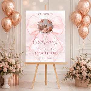 May include: A rectangular welcome sign on a wooden easel featuring a large pink watercolor bow graphic, a circular photo of a child, and text reading: WELCOME TO Caroline's My Bow-tiful 1ST BIRTHDAY JULY 13, 2026, surrounded by rose gold balloons.