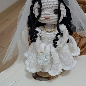 May include: A crocheted doll with black yarn hair, wearing a white dress adorned with small white pearls, a gold-colored necklace, and a sheer white veil. The doll holds a small white fabric flower bouquet and has dark eyes and orange lips.
