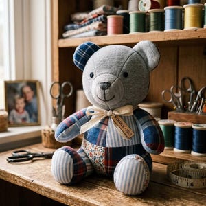 May include: A patchwork teddy bear constructed from grey jersey fabric and various blue, red, and white plaid and floral patterned textiles. The bear features a cream-colored ribbon tied around its neck with a wooden tag engraved with CHARLIE - 2023.