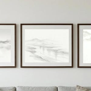 Könnte beinhalten: Ein Set aus drei gerahmten Landschaftsdrucken mit monochromen Tuschezeichnungen von Bergen, Wasser und einer kleinen Hütte. Jeder Druck befindet sich in einem dunkelbraunen Rahmen mit weißem Passepartout und zeigt sanfte Graustufen.