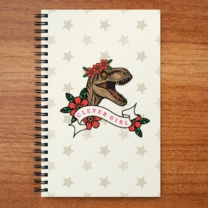 May include: A spiral-bound notebook with an off-white cover featuring a repeating pattern of light gray stars. The center displays a brown dinosaur head wearing a red floral crown, surrounded by red flowers and a white banner with the text CLEVER GIRL.