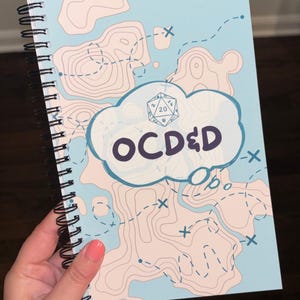 May include: A spiral-bound notebook with a light blue cover featuring a beige topographic map design, dashed path lines, and a central cloud-shaped graphic containing a twenty-sided die icon and the dark purple text OC D&D.