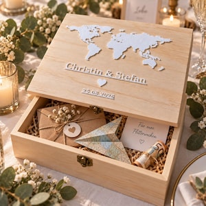 Könnte beinhalten: Eine helle Holzbox mit Klappdeckel, verziert mit einer weißen Weltkarte, den Namen Christin & Stefan, einem Herzsymbol und dem Datum 25.06.2026. Innen liegen ein brauner Umschlag mit Herzanhänger, ein Papierflieger und ein gerollter Geldschein.