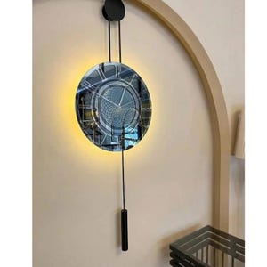 Könnte beinhalten: Eine kreisförmige Wanduhr mit einem dunklen, reflektierenden Zifferblatt, das ein geometrisches Gittermuster und dünne silberne Zeiger aufweist. Die Uhr hängt an zwei schwarzen vertikalen Stangen und hat ein langes, schwarzes Pendel.