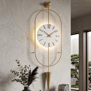 Könnte beinhalten: Eine Wanduhr mit einem runden weißen Zifferblatt und goldenen Ziffern auf 12, 3, 6 und 9 Uhr, umgeben von einem dünnen ovalen Goldrahmen. Die Uhr verfügt über goldene Zeiger, eine Hintergrundbeleuchtung und ein spitzes goldenes Pendel.