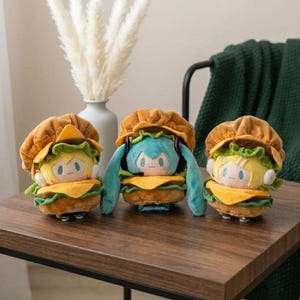 May include: Three plush toys shaped like hamburgers, each featuring a stylized character head nestled between a bun, lettuce, and cheese. The center character has teal hair and headphones, while the two flanking characters have yellow hair.