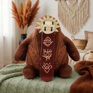 May include: A brown plush toy with a rounded body and arms, featuring a cream-colored sun-shaped face with round glasses. A long, dark red fabric strip hangs from the face, decorated with four distinct gold-colored geometric symbols.