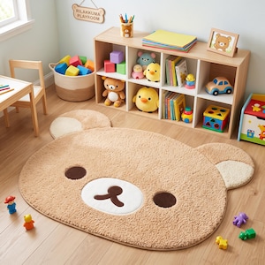 May include: A large, oval, tan-colored plush rug shaped like a bear face with dark brown circular eyes, a white muzzle, and a dark brown nose and mouth. The rug features two rounded ears with lighter tan accents on the top left and right sides.