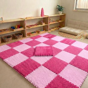 May include: A large, square floor mat featuring a checkered pattern of alternating light pink and dark pink fuzzy squares. The mat is composed of interlocking foam-style tiles, with a single detached square piece resting in the center.