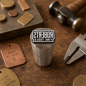 May include: A cylindrical metal stamping tool with the text ROBERTS FINE TOOLS engraved on the face. The tool is surrounded by various copper and brass tags, some stamped with MADE BY HAND and WORK WITH PURPOSE, alongside a hammer and calipers.