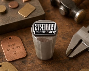 May include: A cylindrical metal stamping tool with the text ROBERTS FINE TOOLS engraved on the face. The tool is surrounded by various copper and brass tags, some stamped with MADE BY HAND and WORK WITH PURPOSE, alongside a hammer and calipers.
