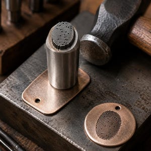 May include: A cylindrical steel metal stamp featuring a raised fingerprint pattern on its face, positioned next to a copper-colored rectangular tag and a circular copper-colored tag, both displaying an impressed fingerprint design.