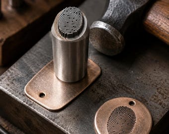 May include: A cylindrical steel metal stamp featuring a raised fingerprint pattern on its face, positioned next to a copper-colored rectangular tag and a circular copper-colored tag, both displaying an impressed fingerprint design.