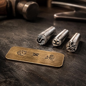 May include: Three cylindrical metal stamping tools with engraved designs on their ends, positioned next to a rectangular brass plate. The plate features three distinct stamped impressions that correspond to the patterns on the metal tools.