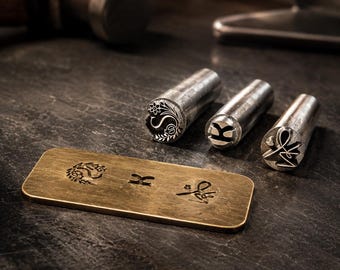 May include: Three cylindrical metal stamping tools with engraved designs on their ends, positioned next to a rectangular brass plate. The plate features three distinct stamped impressions that correspond to the patterns on the metal tools.