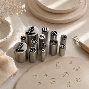 May include: A collection of cylindrical metal stamps with various engraved designs including letters, numbers, symbols, and the text CLÉ CERAMIQUE. The stamps are arranged upright, with some impressions visible on a flat, light-colored clay surface.