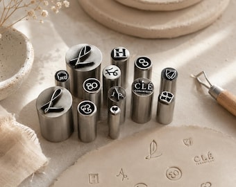May include: A collection of cylindrical metal stamps with various engraved designs including letters, numbers, symbols, and the text CLÉ CERAMIQUE. The stamps are arranged upright, with some impressions visible on a flat, light-colored clay surface.