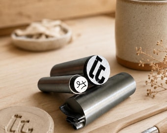 May include: Three cylindrical metal stamps with flat circular ends featuring engraved designs. One stamp displays a stylized letter, another shows a cursive character, and the third is a larger stamp with a bold, geometric letter design.