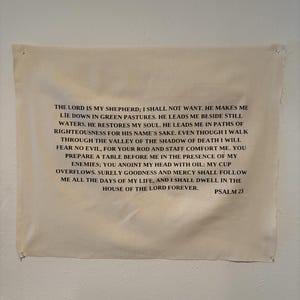 Könnte beinhalten: Ein rechteckiger Wandbehang aus cremefarbenem Stoff mit dem vollständigen Text von Psalm 23 in schwarzen Großbuchstaben. Der Stoff hat rohe, unversäuberte Kanten und ist an den vier Ecken mit kleinen silberfarbenen Metallstiften befestigt.