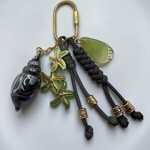 May include: A keychain featuring a gold-toned carabiner, a black and white swirled glass shell charm, three green starfish charms, a translucent green leaf-shaped pendant, and dark gray paracord strands accented with gold-toned metal beads.