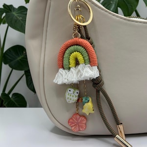 May include: A decorative keychain featuring a multi-colored fiber rainbow in shades of coral, green, and yellow with white fringe. Attached are three small charms: a white owl, a yellow bird, and a coral flower, all suspended by gold chains.