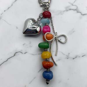 May include: A silver-toned keychain featuring a heart-shaped charm and a vertical strand of multi-colored, polished stones in red, light blue, pink, orange, green, yellow, rust, and blue, accented with a silver metal bow and small spacers.