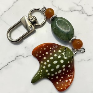May include: A keychain featuring a ceramic stingray pendant with a green and orange gradient and white spots. The pendant is attached to a silver-tone metal lobster clasp by a strand of beads, including one large oval green stone and two small orange beads.