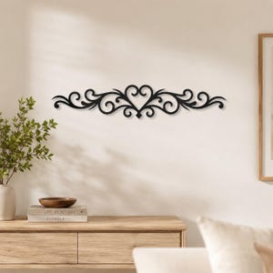 Peut inclure: Une pièce décorative murale noire présentant un design horizontal symétrique avec des volutes complexes et un motif central en forme de cœur. L'objet est fabriqué dans un matériau plat et affiche un motif continu et fluide.