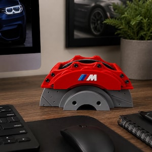 May include: A red automotive brake caliper model featuring the M logo in blue, light blue, and white, mounted on a gray ventilated brake rotor base. The object is constructed with visible black screws and has a textured, realistic design.
