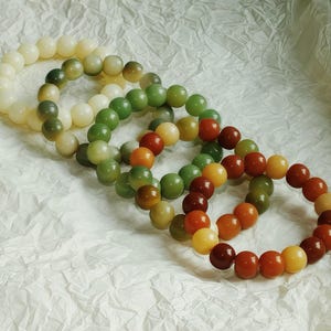 May include: Three circular bracelets made of smooth, polished spherical beads. One bracelet features cream-colored beads, another consists of various shades of green, and the third displays a mix of warm amber, orange, and deep reddish-brown tones.