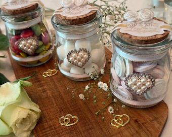May include: Three clear glass jars with cork lids, each topped with two white fabric roses. The jars are filled with colorful candies, white spheres, and white discs. Each jar features a white ribbon and a heart-shaped pearl embellishment.
