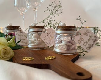 May include: Three small glass jars with cork stoppers, filled with white candies and pearl accents. Each jar features a white ribbon and a decorative tag with the names Susan, John, and Samantha written in gold script, placed on a wooden board.