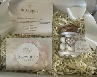 May include: A gift box containing a glass jar filled with white candies, topped with a cork lid and a heart-shaped pearl embellishment. A white tag labeled Susan is attached. Includes a Rosemere Studio card and a packet of dried flower petals.