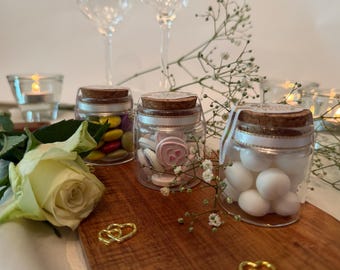 May include: Three small clear glass jars with cork stoppers, each filled with different candies. One jar contains multi-colored candies, another holds pink heart-shaped candies, and the third is filled with white round candies.