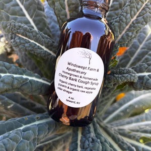 Puede incluir: Una botella de 4 onzas de jarabe para la tos de corteza de cerezo de Windswept Farm & Apothecary. La botella es de vidrio marrón con una etiqueta blanca que dice "Homegrown & Homemade" y "Organic cherry bark, vegetable glycerin & organic raw apple cider vinegar".