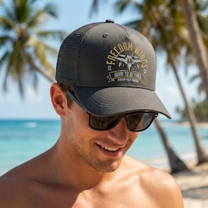 May include: A charcoal gray trucker hat with a mesh back and a curved brim. The front panel features a graphic of a fighter jet with the text "FREEDOM WINGS," "ESTD 2026," "BORN TO BE FREE," and "KEEP FLY HIGH" printed in yellow and white.