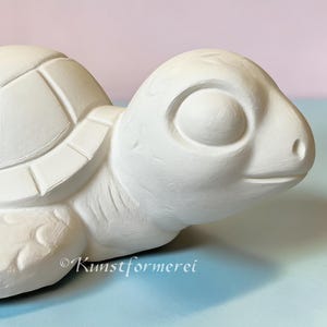 May include: A white ceramic turtle figurine with a smooth, matte finish. The turtle features a rounded shell with clearly defined geometric plate patterns, large eyes, and textured flippers. The text Kunstformerei is visible on the side.