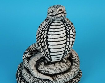 May include: A white and black cobra figurine coiled in a circular base. The snake is depicted in an upright, alert posture with its hood flared, showing detailed scale textures and dark shading between the scales on its body and head.