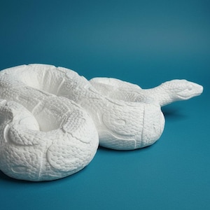 Peut inclure: Une sculpture de serpent blanc enroulé présentant une surface texturée imitant des écailles sur tout le corps. La figurine est représentée dans une position de repos, en boucle, avec la tête légèrement relevée et orientée vers la droite.