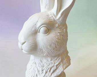 May include: A white matte rabbit bust sculpture featuring detailed fur texture, long upright ears, and a prominent eye. The rabbit head is mounted on a smooth, circular pedestal base. The text Kunstformerei is visible at the bottom of the image.