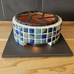 Könnte beinhalten: Ein runder Kerzenhalter aus Keramik mit einem Mosaik-Außenbereich aus kleinen, glänzenden blauen und salbeigrünen Fliesen. Die Oberseite ist mit einem schwarzen Metallgitter versehen und das Innere zeigt eine Basis aus Terrakotta auf einer schwarzen Fliese.
