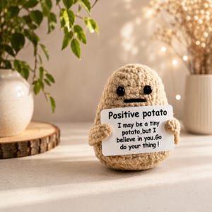 Könnte beinhalten: Eine gehäkelte Kartoffelfigur in Beige mit schwarzen Perlenaugen und einem flachen Mund. Sie hält ein weißes rechteckiges Schild mit der Aufschrift: Positive potato. I may be a tiny potato, but I believe in you. Go do your thing! Die Figur ist texturiert.