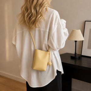 May include: A light yellow crossbody bag with a smooth, vertical rectangular shape and a thin shoulder strap. The bag features a decorative knotted detail on the side, hanging from the strap attachment point, and has a clean, minimalist exterior.