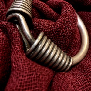 Può includere: Un anello in metallo color argento caratterizzato da due distinte sezioni a molla avvolte e integrate nella fascia circolare. Il metallo presenta una finitura opaca liscia con una visibile ossidazione scura nelle fessure delle spire, creando un contrasto con il tessuto bordeaux.
